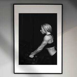 May include: Black and white photograph of a woman lifting weights in a gym. She is wearing a black sports bra and black shorts. The photo is framed in a black frame.