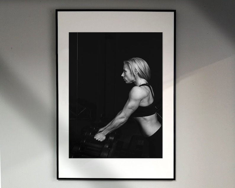 May include: Black and white photograph of a woman lifting weights in a gym. She is wearing a black sports bra and black shorts. The photo is framed in a black frame.