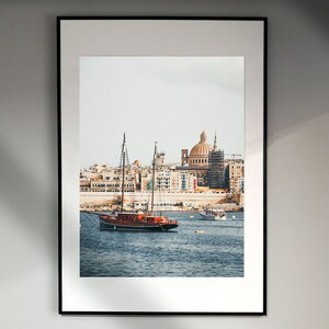 May include: A framed photograph of a red sailboat docked in a harbor with a city skyline in the background. The city skyline features a large domed building.
