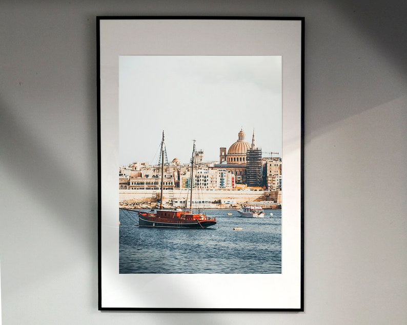 May include: A framed photograph of a red sailboat docked in a harbor with a city skyline in the background. The city skyline features a large domed building.