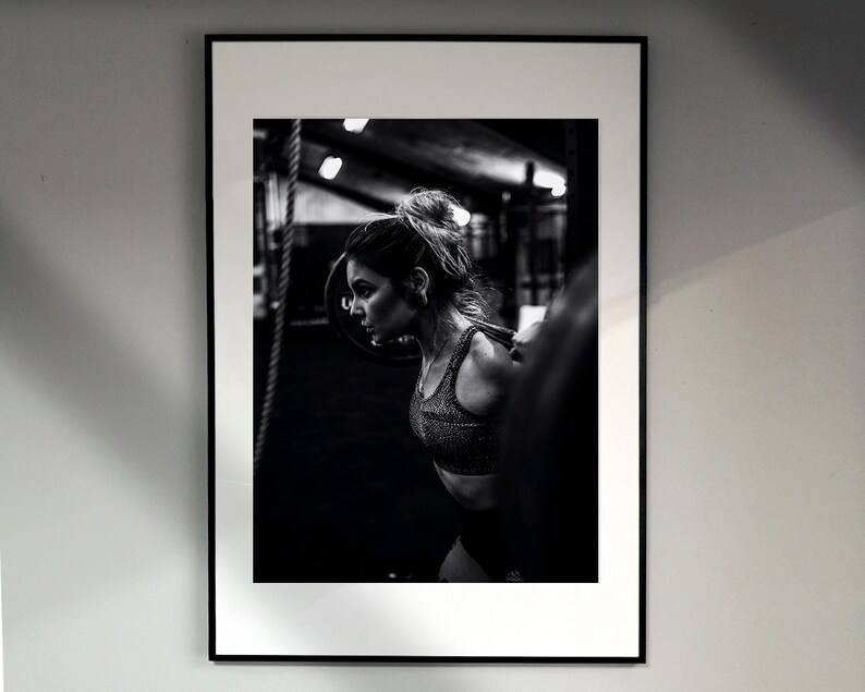 May include: Black and white photo of a woman lifting weights in a gym. She is wearing a black sports bra and has her hair pulled back in a ponytail.
