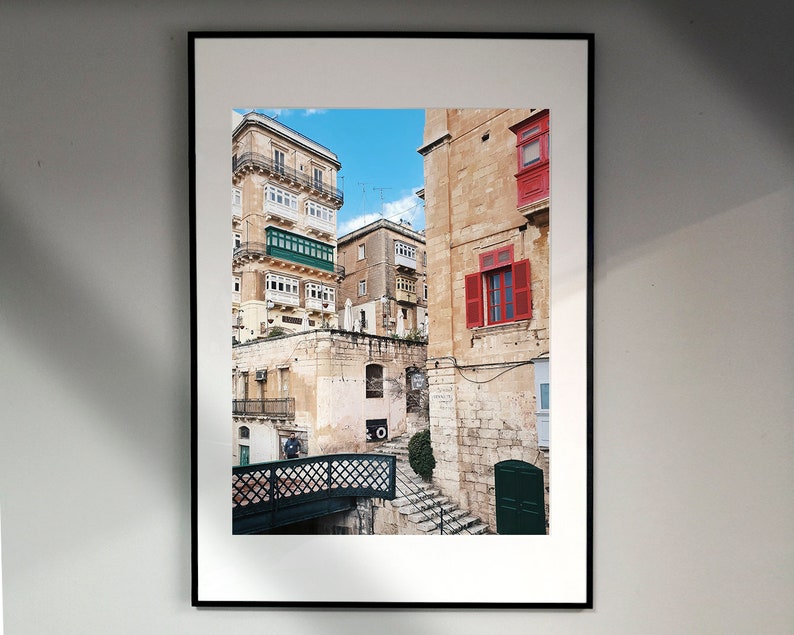 May include: A framed photograph of a narrow street in a European city. The buildings are made of stone and have red and green window shutters. A small bridge crosses the street in the foreground.