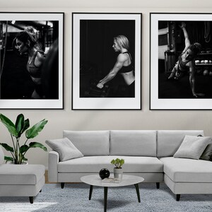 May include: Three black and white framed photographs of a woman working out in a gym. The photos are hung on a wall above a gray sectional sofa and a coffee table.