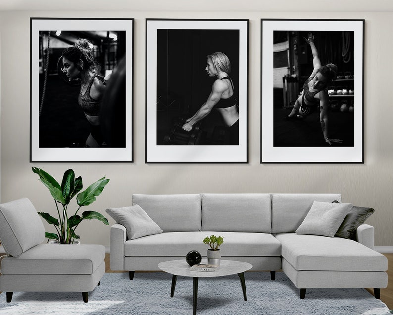 May include: Three black and white framed photographs of a woman working out in a gym. The photos are hung on a wall above a gray sectional sofa and a coffee table.