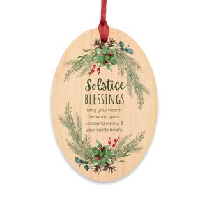 以下が含まれることがあります： 「Solstice Blessings」の文字と赤いリボンが付いた、楕円形の木製クリスマスオーナメント。オーナメントは、松葉、ヒイラギ、赤い実の緑色のリースで飾られています。テキストは「May your hearth be warm, your company merry, & your spirits bright.」と書かれています。