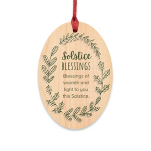 以下が含まれることがあります： 「Solstice Blessings」の文字と緑の葉のデザインが施された木製の楕円形のオーナメント。オーナメントには吊り下げ用の赤いリボンが付いています。テキストは「この冬至にあなたに暖かさと光の祝福を」と書かれています。