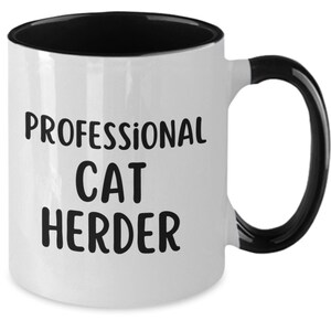 May include: White ceramic coffee mug with a black rim and handle. The mug has black text that reads "Professional Cat Herder".