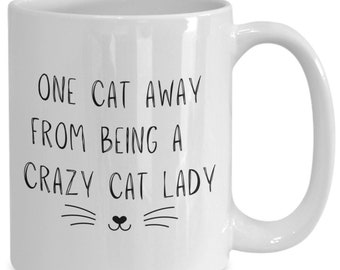 May include: White ceramic coffee mug with black text that reads "One cat away from being a crazy cat lady" and a black cat face with whiskers.