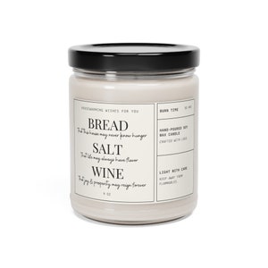 May include: A white soy wax candle in a glass jar with a black lid. The candle has a label that reads "Housewarming Wishes for You" and features the text "Bread, Salt, Wine" with a saying about each. The candle is 9 oz and has a burn time of 50 hours.