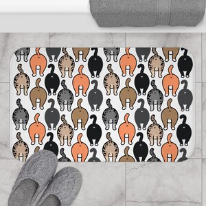 Può includere: Un tappetino da bagno rettangolare con un motivo ripetuto di sederi di gatto in vari colori, tra cui arancione, nero, marrone e grigio, su sfondo bianco. Pantofole grigie in primo piano.