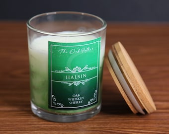 May include: A glass candle with a green and white interior, labeled "Halsin" with "Oak Whiskey Sherry." The candle sits next to its wooden lid. The label is green with white text and decorative elements. The candle is in a clear glass container.