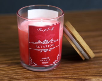 May include: A red glass candle jar with a wooden lid. The label reads "The pale of Astarion Amber Vanilla Musk".