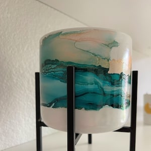 May include: A white ceramic planter with a teal and gold abstract design sits on a black metal stand.