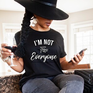May include: A woman wearing a black t-shirt with the text "I'm not for everyone" in white lettering. She is holding a glass of red wine and a black smartphone.