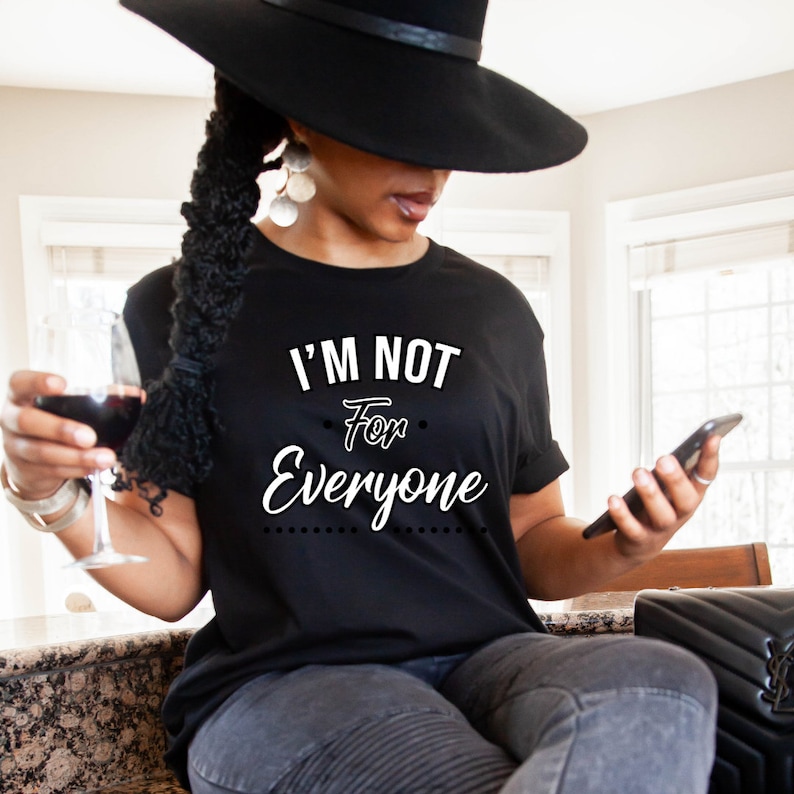 May include: A woman wearing a black t-shirt with the text "I'm not for everyone" in white lettering. She is holding a glass of red wine and a black smartphone.