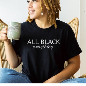 May include: A person wearing a black t-shirt with the text "ALL BLACK everything" in white lettering. The person is holding a green mug with a dragonfly design.