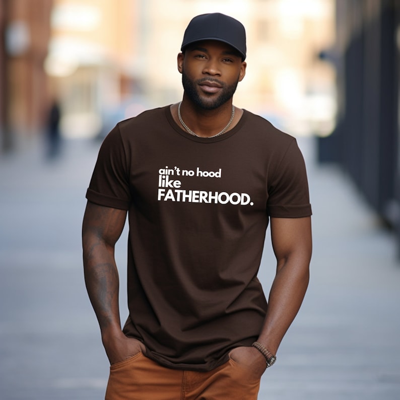 May include: A brown t-shirt with white text that reads "ain't no hood like FATHERHOOD."