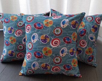 May include: Four teal blue throw pillows with a pattern of colorful umbrellas and waves. The pillows are arranged in a row on a dark wood floor.