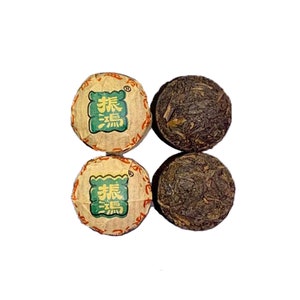 May include: Four round tea cakes, two are wrapped in brown paper with green and orange Chinese characters that read "flavor good", and two are unwrapped and show the dark brown compressed tea leaves.