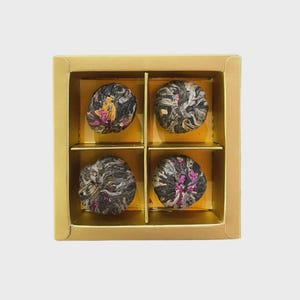 May include: A gold box containing four tea flower balls. Each ball is a tightly wound bundle of dark leaves with visible pink and yellow flower petals. The box has four compartments, each holding a tea ball.