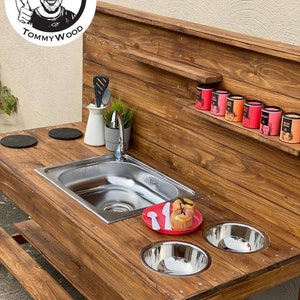 Könnte beinhalten: Holzspeiseküche für den Außenbereich mit einem Edelstahlspülbecken, zwei Schüsseln, einem roten Teller und zwei Utensilien. Die Küche hat eine Holzrückwand mit einem Regal und Haken. Die Küche ist aus Naturholz mit einer dunkelbraunen Beize.