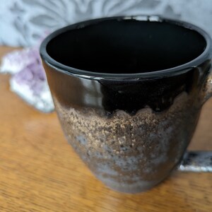 Könnte beinhalten: Eine schwarze Keramiktasse mit metallischer Gold- und Bronzeglasur. Die Tasse hat eine abgerundete Form und einen Henkel. Das Innere der Tasse ist schwarz. Die Tasse steht auf einer Holzoberfläche.