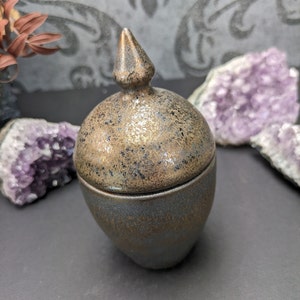 Könnte beinhalten: Eine kleine, keramische Dose mit einem spitzen Deckel. Die Dose ist dunkelgrau mit einer gesprenkelten, goldenen Glasur. Der Deckel ist in einem dunkleren Grauton mit einer goldenen Glasur.