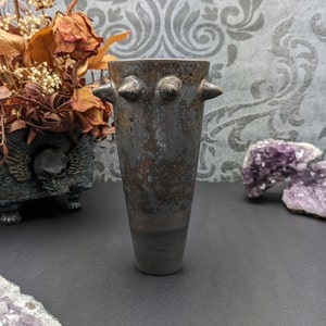 Könnte beinhalten: Eine hohe, dunkle Keramikvase mit strukturierter Oberfläche und zwei Paaren von hervorstehenden, konischen Formen nahe der Oberseite. Die Vase hat eine dunkle, gesprenkelte Oberfläche mit grünem Sockel. Die Vase dient wahrscheinlich dekorativen Zwecken.