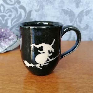 Könnte beinhalten: Schwarze Keramiktasse mit einer weißen Silhouette einer Hexe, die auf einem Besen fliegt.