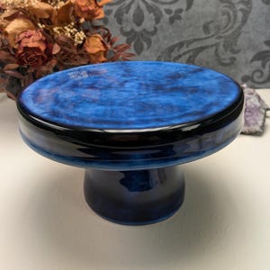 Könnte beinhalten: Ein Keramik-Kuchenständer mit tiefblauer Glasur. Der Ständer hat eine runde Oberseite und einen Sockel. Die Oberseite hat einen Durchmesser von etwa 20 cm. Der Ständer ist ein dekoratives Stück zum Präsentieren von Desserts.