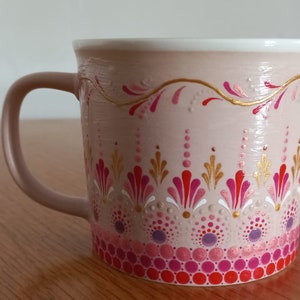Puede incluir: Una taza de cerámica color rubor con borde blanco y asa a juego. La taza está decorada con un patrón repetitivo de diseños florales rosas, rojos y dorados. El diseño está pintado a mano y presenta un patrón de puntos.
