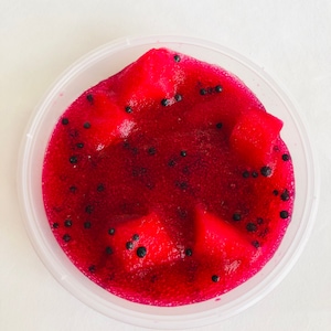 May include: A clear plastic container filled with red slime with black specks. The slime is shaped like a dragon fruit.