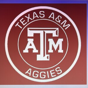 Puede incluir: Un círculo blanco con las palabras "Texas A&M Aggies" alrededor del exterior. Dentro del círculo hay una "T" blanca con una "A" blanca y una "M" a cada lado de la "T". El fondo es marrón.