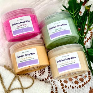 Op de afbeelding: Vier potten Sabrina Soap Bliss whipped foaming sugar scrub in roze, groen, oranje en beige. Elke pot heeft een doorzichtige deksel en een etiket met de productnaam en geur. De potten staan op een wit en bruin patroon.
