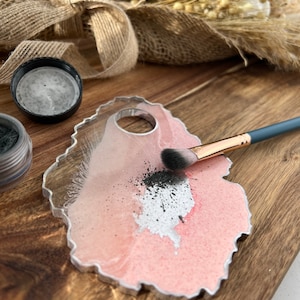 May include: A pink and white resin makeup palette with a silver rim and a round indentation in the centre. Black eyeshadow is spread on the palette with a small makeup brush.
