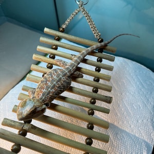 Puede incluir: Un lagarto barbudo marr&oacute;n y blanco trepando por una escalera de bamb&uacute; colgante con cuentas de madera marr&oacute;n. La escalera est&aacute; suspendida del techo con una ventosa y una cuerda.