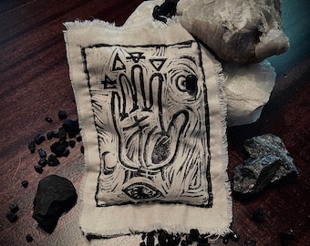 May include: A black and white hand print design on a beige fabric pouch. The pouch is surrounded by black stones.