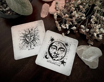 May include: Two white cards with black ink illustrations of the sun and moon. The sun card says "Action" and the moon card says "Introspection". The cards are surrounded by white crystals, dried flowers, and a lit candle in a glass jar.