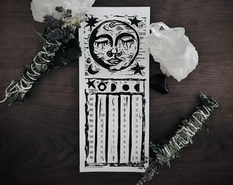 May include: A black and white print of a moon with a face and stars. The print is on a white background and has a calendar for the year 2022. The calendar is divided into months and days. The print is surrounded by white crystals and sage bundles.