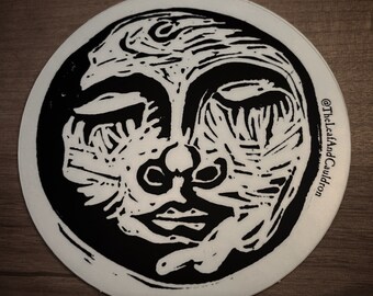 May include: Black and white circular sticker with a stylized face design. The face has a closed eye, a nose, and a mouth. The text "@TheLeafAndCauldron" is printed on the right side of the sticker.