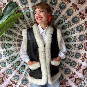 Op de afbeelding: Zwart leren vest met witte schapenvacht afwerking en zakken. Het vest heeft een knoopsluiting en een witte schapenvacht kraag. Het model draagt een wit overhemd en een blauwe jeans.