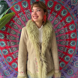 Peut inclure: Veste en daim beige avec col et bordure en fausse fourrure vert clair. La veste présente des broderies florales sur les poignets et le devant. Une pièce d'inspiration vintage avec une esthétique bohème.