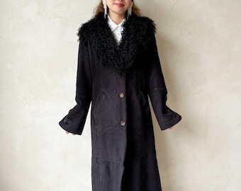 May include: A long black coat featuring a large, textured black faux fur collar, long sleeves with buttoned cuffs, and a front button closure. The coat is worn over a white collared shirt and blue denim jeans, creating a layered look.