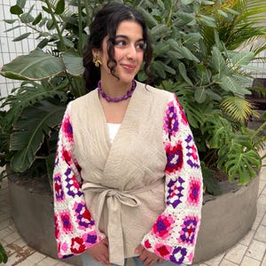 Op de afbeelding: Een beige gewatteerde jas met een strik in de taille en gehaakte mouwen. De mouwen hebben een kleurrijk granny square patroon in roze, rood en paars. De jas wordt gedragen met een blauwe spijkerbroek.