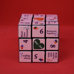 May include: A pink cube with black edges and numbers 1 through 9 on each side. Each side also has a different activity suggestion, such as "Music Lists Created", "Game Nights", "Books read to each other", "May-Jun", and "Workouts Together".