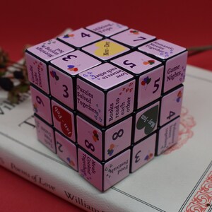 May include: A pink and black cube with numbers and phrases on each side. The phrases include "Game Nights", "Books to read to each other", "Puzzles Solved Together", "Candle Light Dinner", "Staying in Mornings", and "Cooked Meals Together".