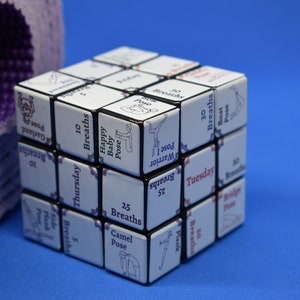 May include: A black and white yoga cube with different yoga poses and breath counts printed on each side. The cube is next to a rolled up purple yoga mat.