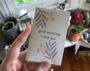 Può includere: Un piccolo biglietto bianco con una foglia viola e stelle arancioni. Il biglietto dice "good morning, i love you".