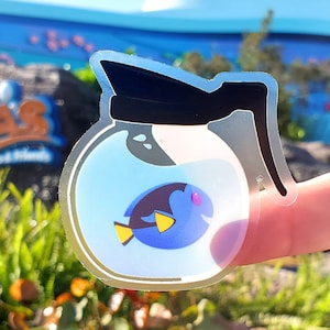 May include: A clear sticker featuring a cartoon image of Dory from Finding Nemo inside a coffee pot. The sticker has a black handle and lid, with a blue and yellow fish design. The background is a blurred outdoor scene.