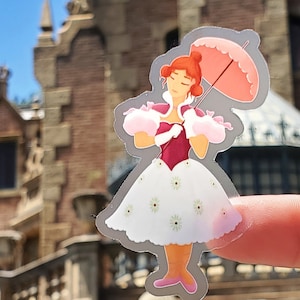 May include: A sticker featuring a cartoon woman in a white dress with small green flowers, a pink parasol, and a burgundy bodice. The woman has red hair and is wearing pink shoes. The sticker is held up in front of a blurred building.